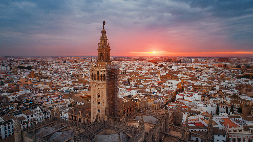 An aerial view of Seville Cathedral at sunrise, with soft golden light illuminating its intricate architecture and surrounding rooftops, casting long shadows across the cityscape.