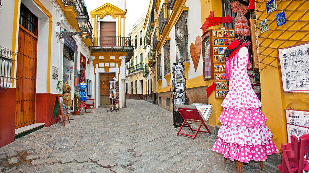 An inviting alleyway in Seville, Spain, lined with charming shops. A colorful dress is displayed on a hanger, adding a touch of vibrancy to the scene.
