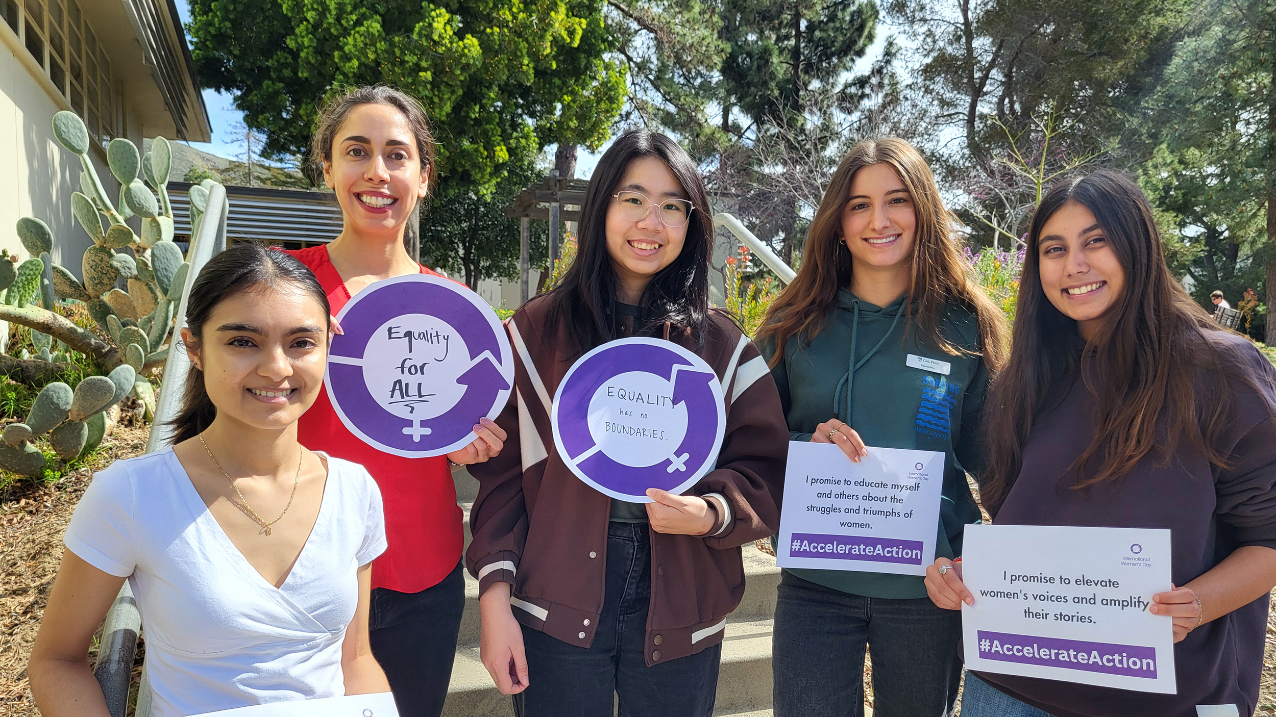 A group of five young women stand outdoors on a sunny day, smiling and holding signs advocating for gender equality. The signs include messages like "Equality for All," "Equality has no boundaries," and pledges to celebrate women's achievements, educate others about women's struggles and triumphs, and amplify women's voices. The signs also feature the hashtag #AccelerateAction.