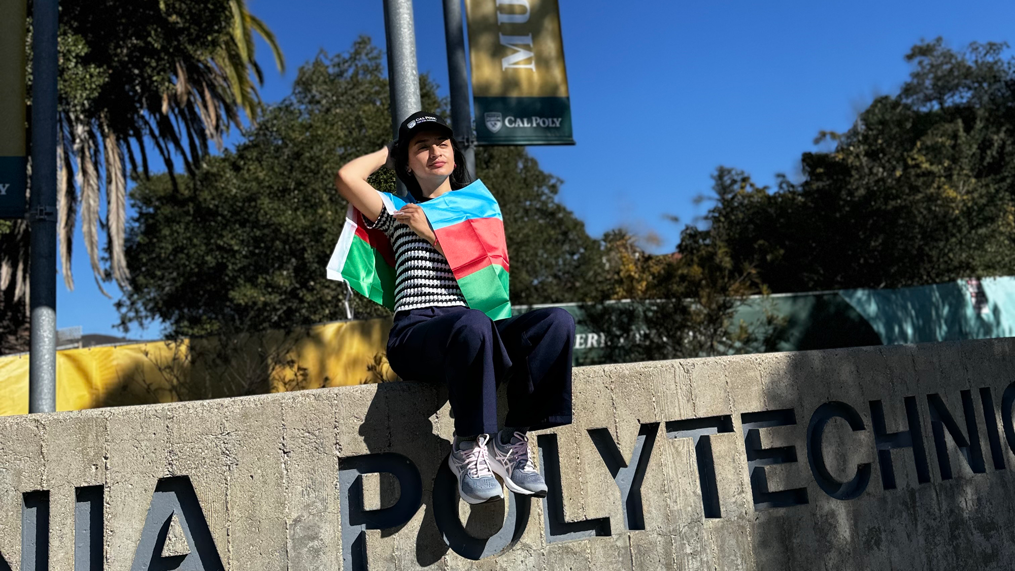 Aynur A young woman sits atop a concrete wall with "CALIFORNIA POLYTECHNIC" engraved on it, wearing a Cal Poly hat and draped in the Azerbaijan flag. She poses confidently under a clear blue sky, with palm trees and campus banners in the background.