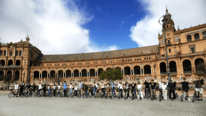 A large group of students poses with bicycles in front of the iconic Plaza de España in Seville, Spain. They are lined up in a row with arms raised in celebration, beneath the building's ornate arches and colorful tilework. The sky above is partly cloudy with a bright patch of blue.