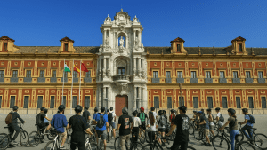 A group of students on bicycles gathers in front of the ornate Baroque entrance of the Palacio de San Telmo in Seville, Spain. They face the historic red-and-gold building, listening attentively, likely during a guided tour. Three flags fly in front of the palace under a clear blue sky.