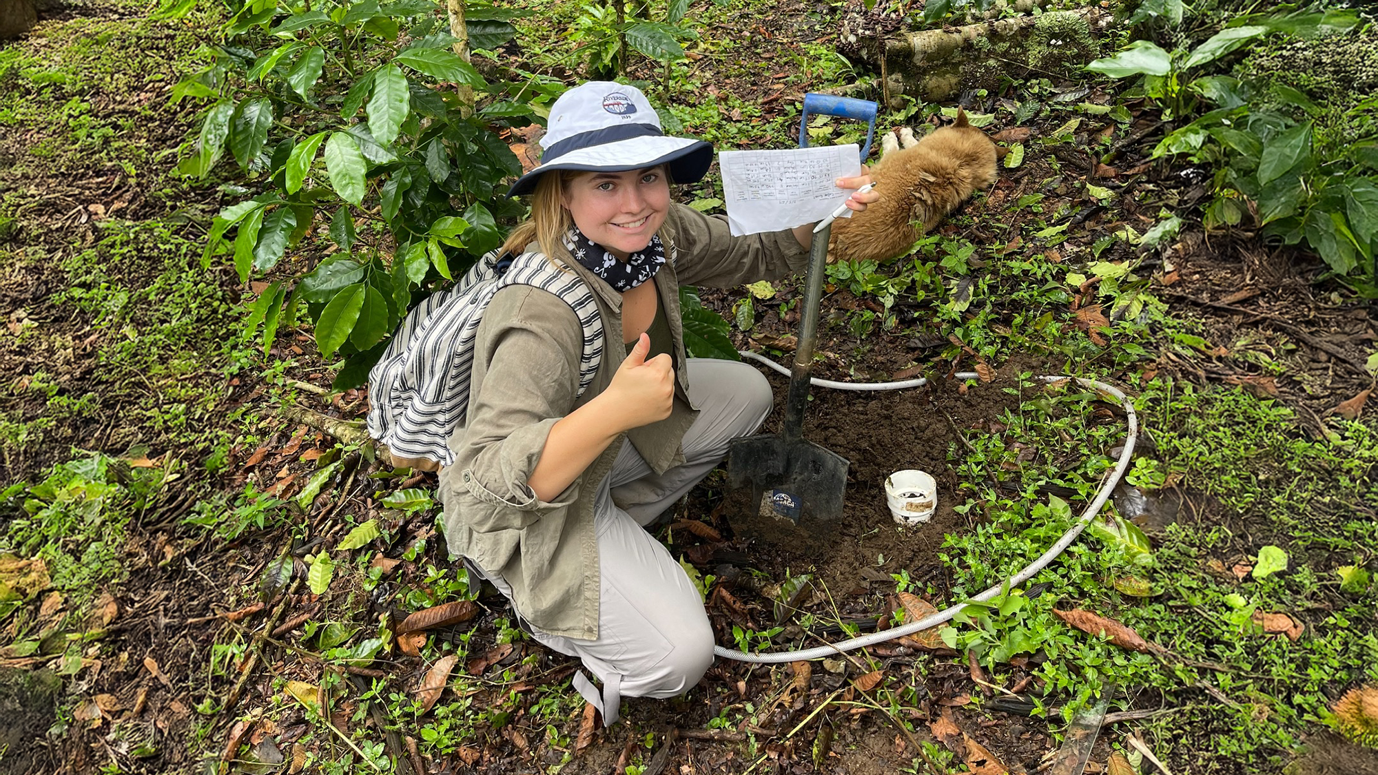 Chelsea Flood 3_CP in Costa Rica A student smiles and gives a thumbs-up while conducting field research in a lush, green forest. Wearing a wide-brimmed hat and outdoor gear, she kneels beside a shovel and a small soil monitoring setup, holding a sheet of data collection notes. A dog lies resting in the background among the plants, adding to the relaxed and immersive nature of the scene.