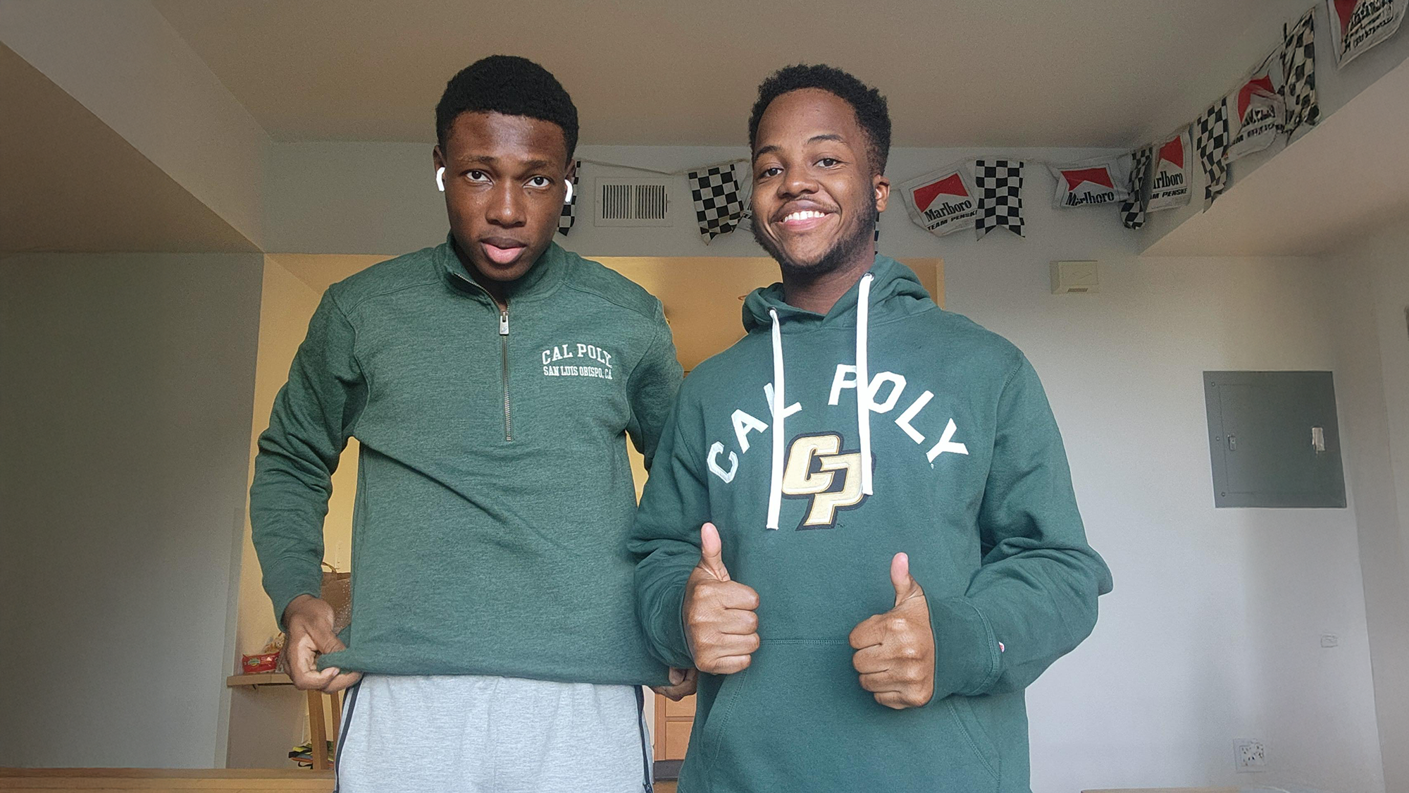 Daryll Two young men pose indoors wearing green Cal Poly San Luis Obispo sweatshirts. One holds the hem of his pullover while the other gives two thumbs up and smiles. Racing-themed flags with Marlboro branding hang along the ceiling in the background.