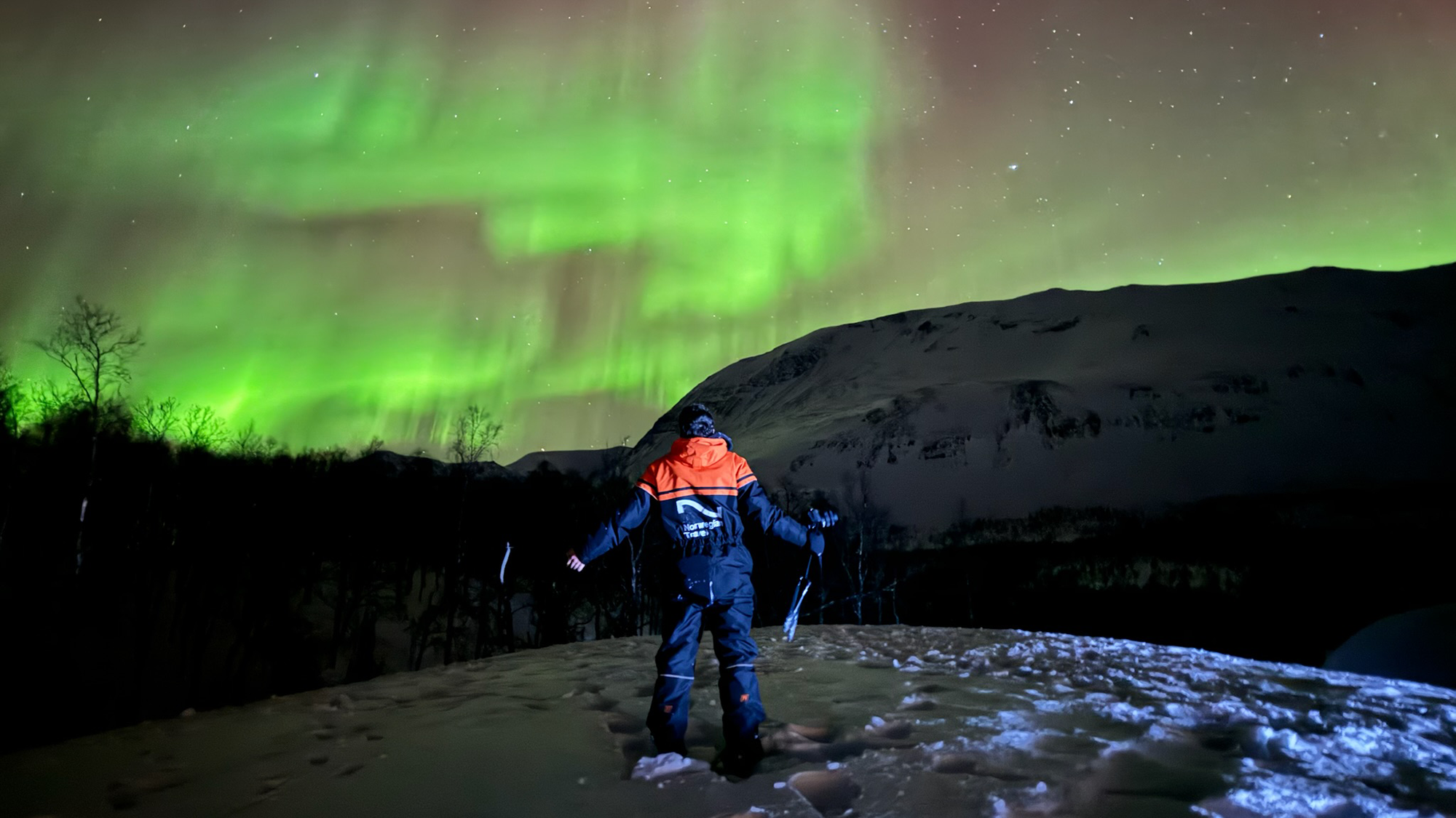 IMG_1427_Oliver Sabin (3) A person in a snowsuit stands on a snowy hill at night, gazing up at a stunning display of the Northern Lights. Vibrant green auroras swirl across the star-filled sky above dark trees and a snow-covered mountain, capturing a moment of awe and wonder in a remote winter landscape.