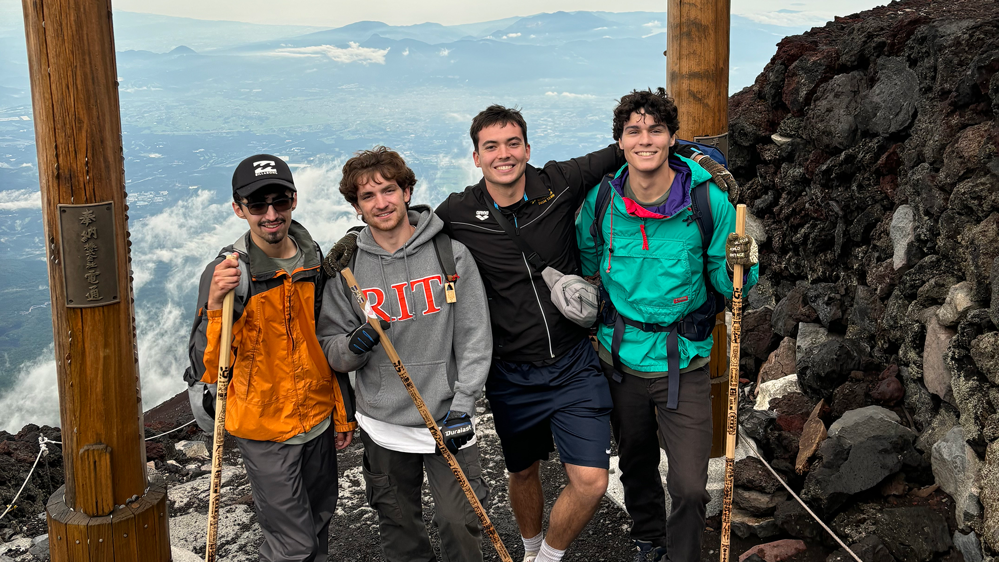 IMG_2845_Kaan Four young men smile while posing at a mountain summit, surrounded by clouds and rocky terrain. They are dressed in hiking gear and holding wooden walking sticks, standing between two large wooden posts with Japanese inscriptions. A scenic valley is visible far below in the background.