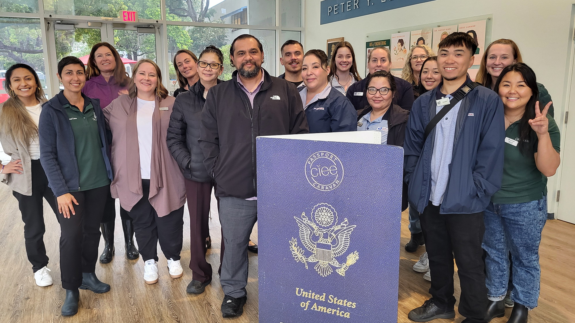 Passport-2025 A diverse group of staff and volunteers pose together indoors around a large cardboard cutout of a U.S. passport displaying the “CIEE Passport Caravan” logo. They are smiling and wearing name badges, suggesting a campus or community event related to international travel or study abroad opportunities. The setting appears to be a university building, with posters and signage visible in the background.