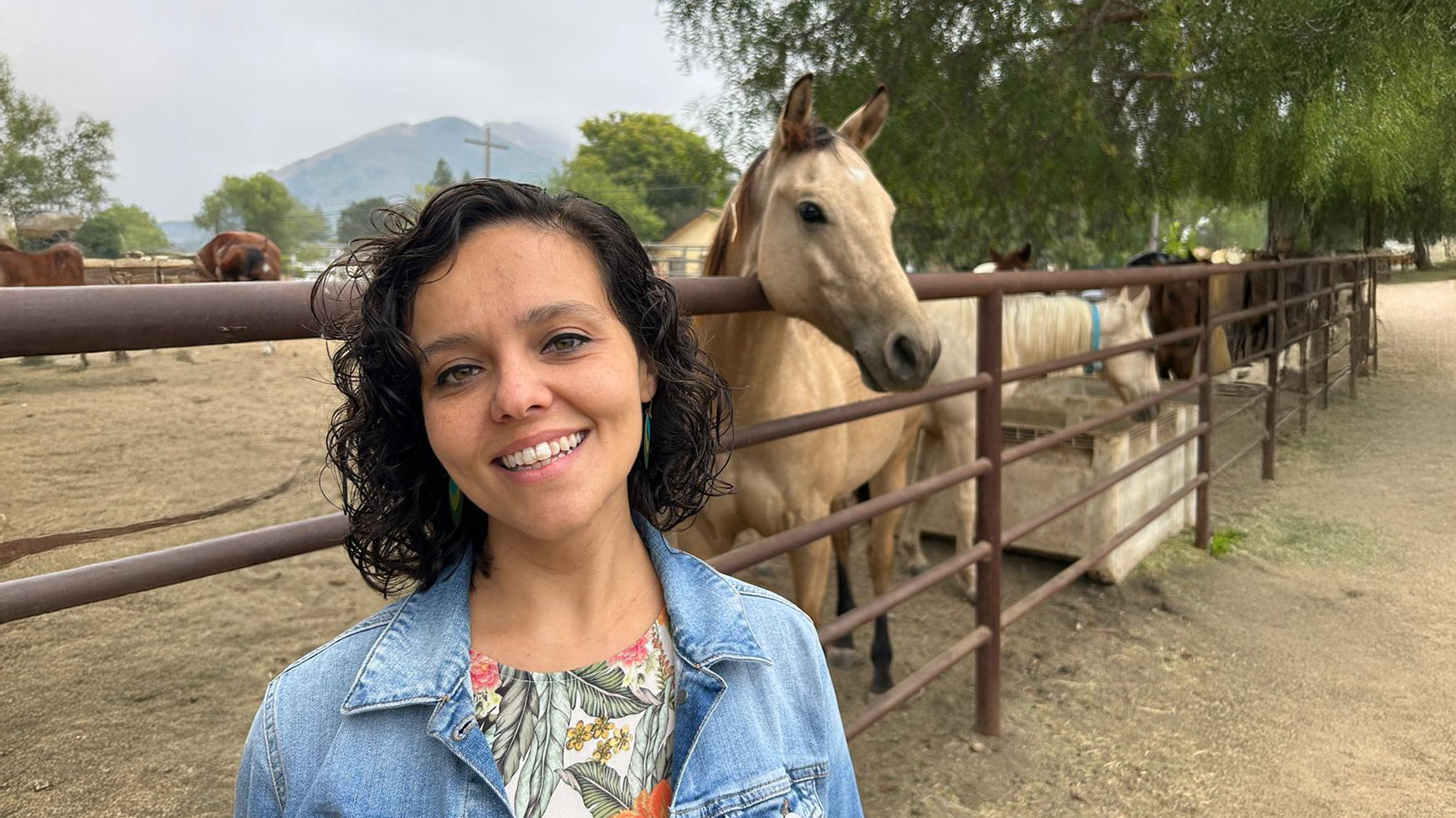 Sinem A woman smiles in front of a fenced horse enclosure, with several horses visible behind her. She is wearing a denim jacket over a floral top, and the background features a dirt paddock, trees, and a misty hill in the distance, creating a peaceful rural setting.