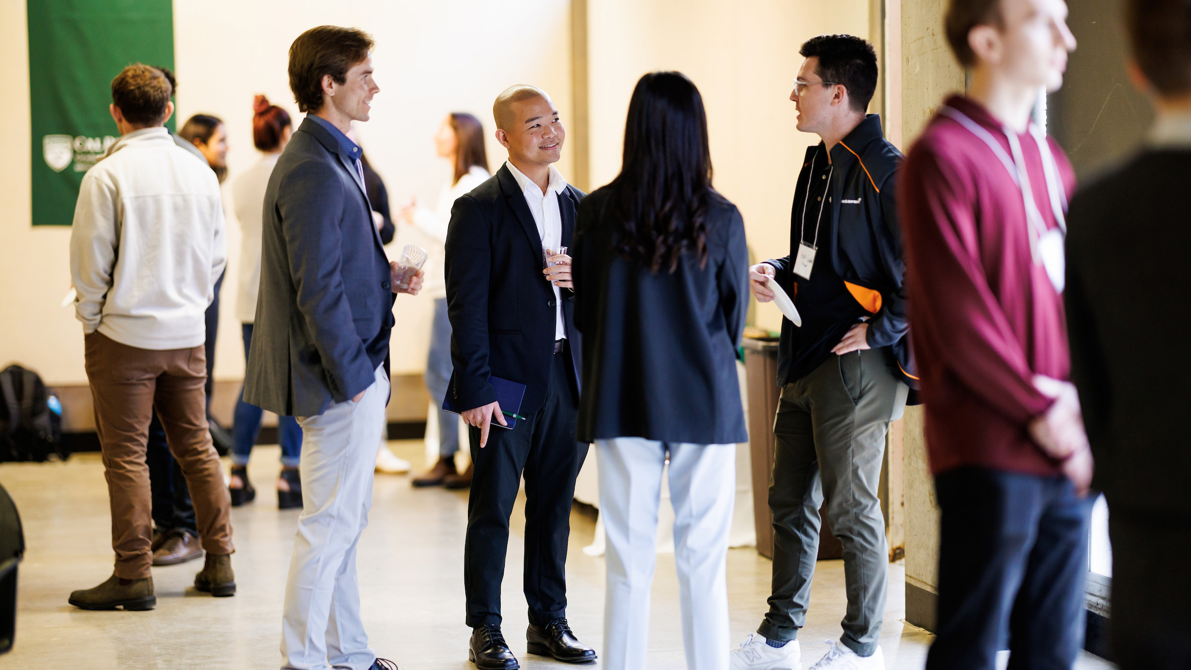 Symposium Attendees at a professional symposium engage in conversation in a well-lit hallway. Small groups of people are seen networking, many wearing name badges. A green Cal Poly banner is visible in the background, indicating a university-hosted event.
