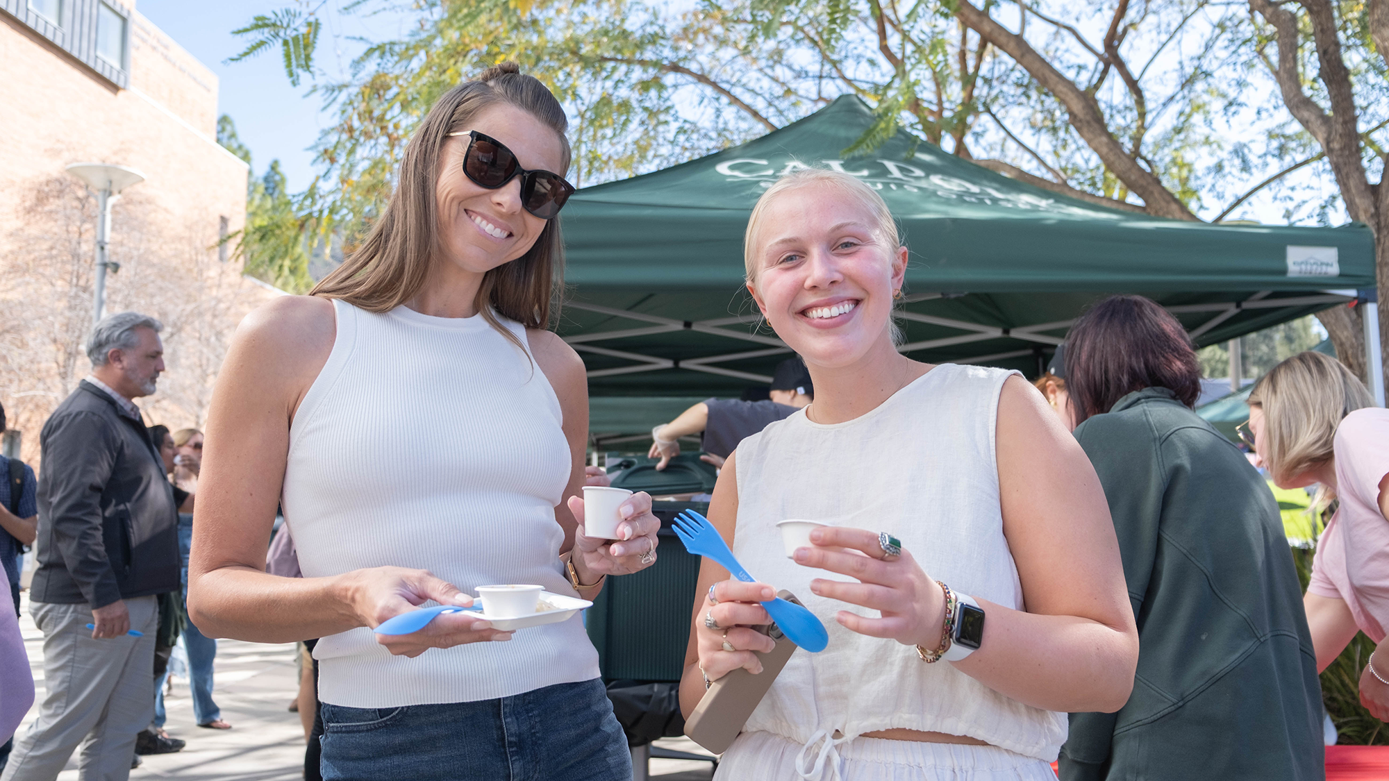 TOTW-2025 Two women smile while enjoying samples of food at an outdoor event, holding small cups and blue utensils. They stand in front of a green Cal Poly-branded canopy, with other attendees and trees visible in the background. The atmosphere is casual and cheerful, likely part of a campus food or cultural celebration.