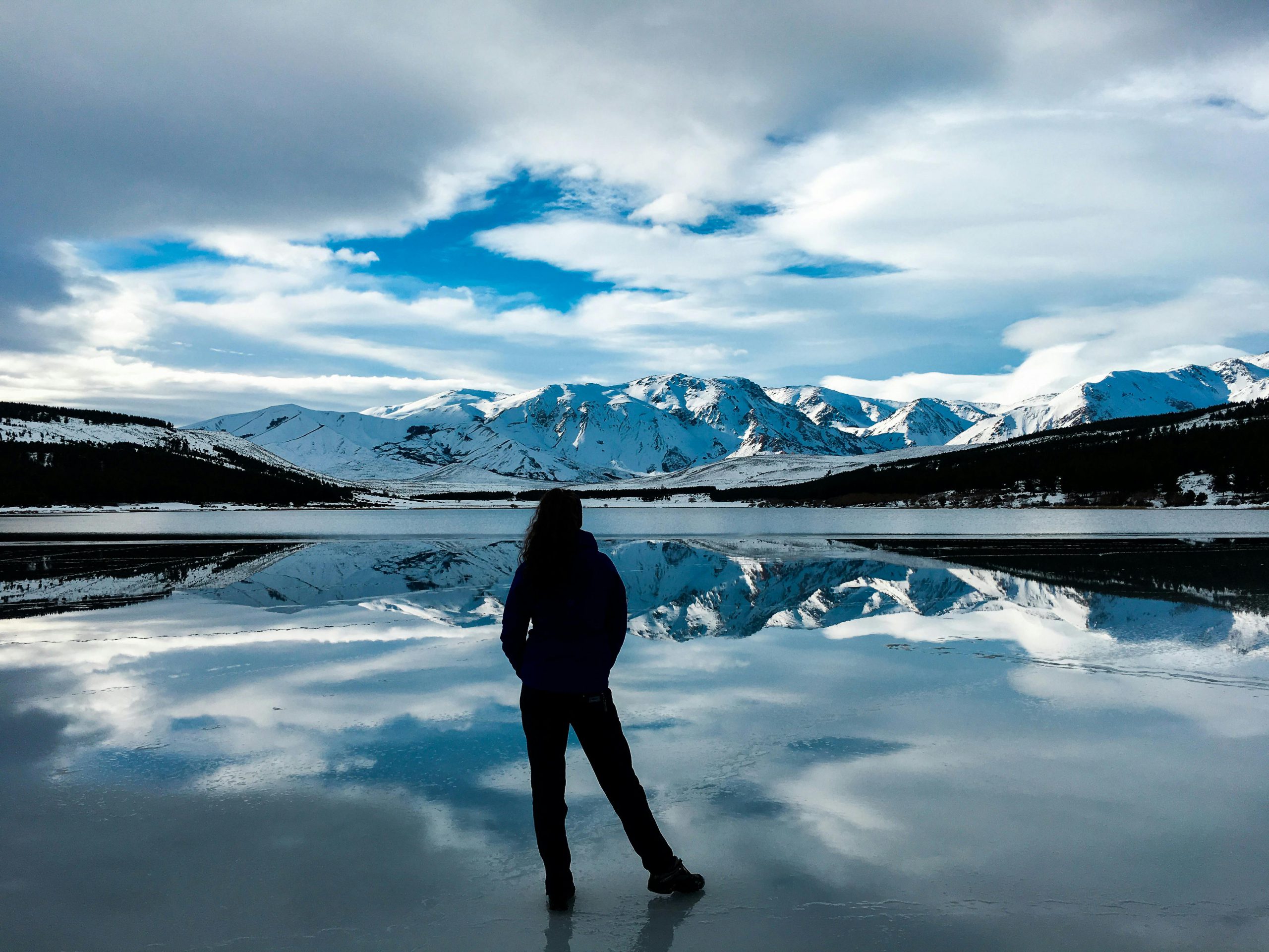 Person standing by a reflective lake with snow-capped mountains in Patagonia, Argentina.