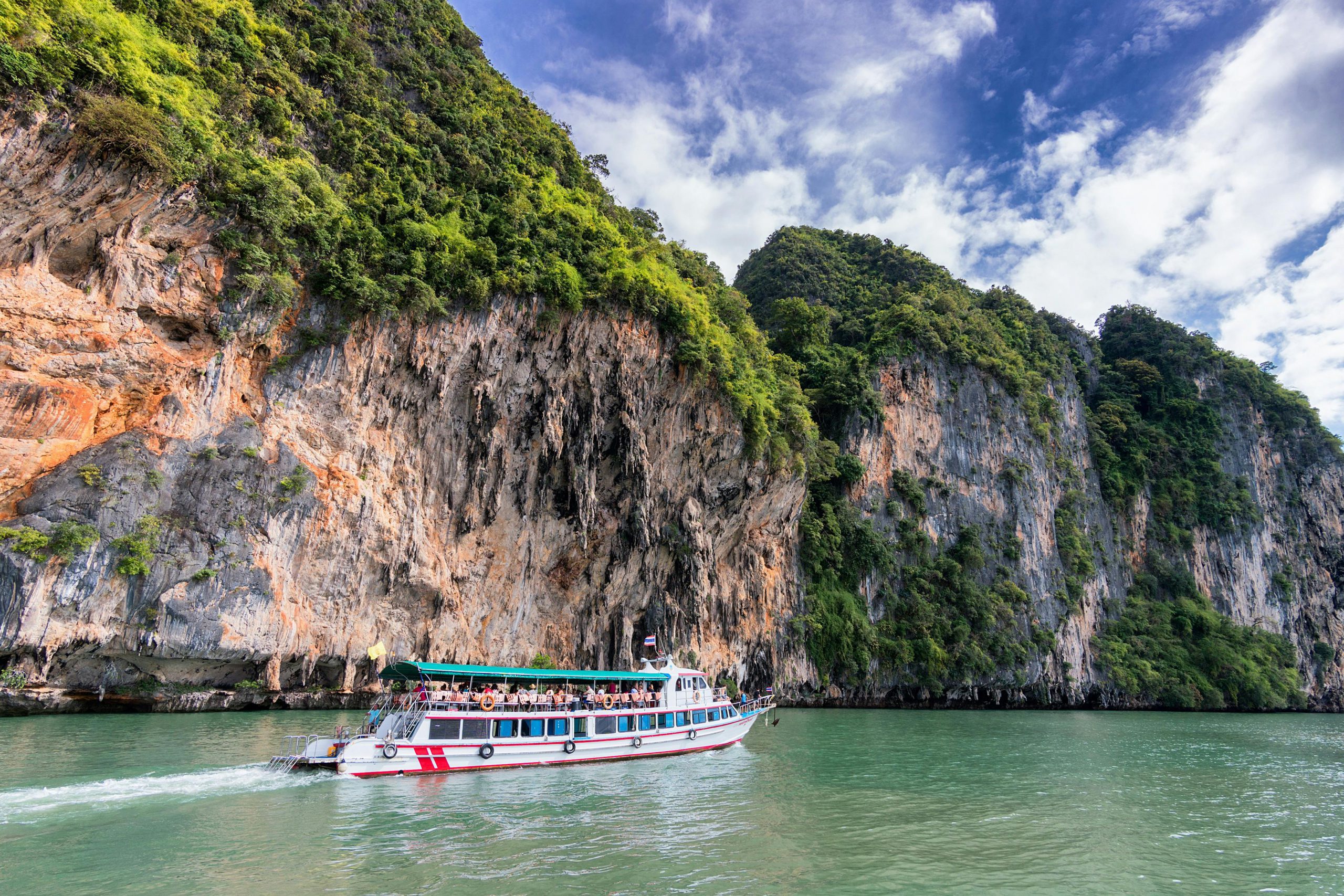 Tour boat cruising past towering limestone cliffs covered in greenery in Thailand.