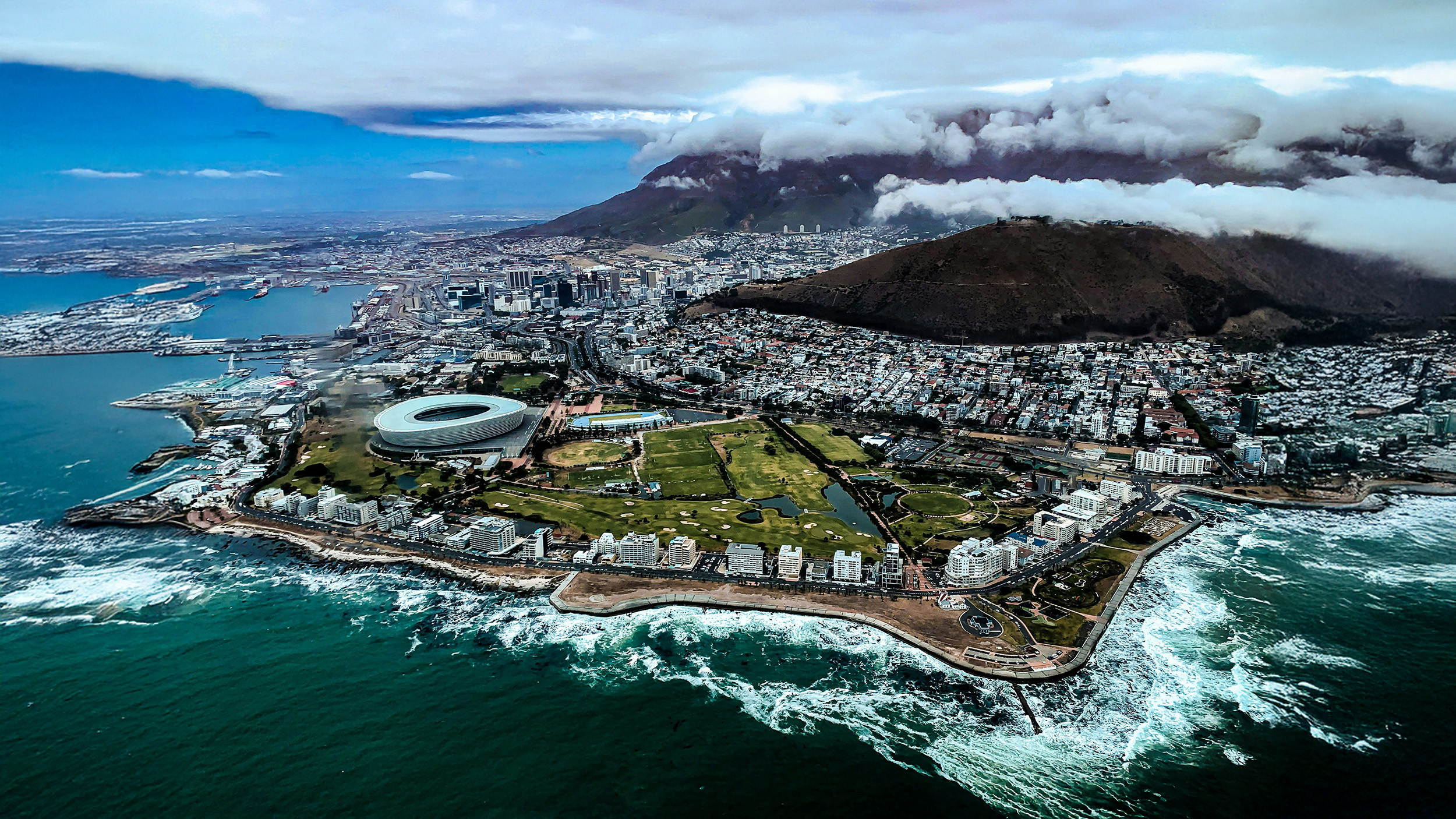An aerial view of Cape Town, South Africa, showing the coastline, Cape Town Stadium, the cityscape, and Table Mountain partially covered by clouds in the background.