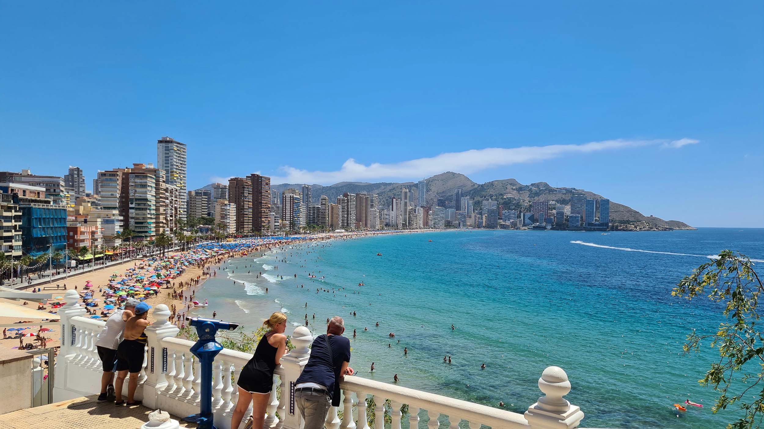 People standing at a white balcony overlooking Benidorm’s beachfront, with colorful umbrellas, swimmers, and tall resort buildings lining the Mediterranean coast under a clear blue sky.