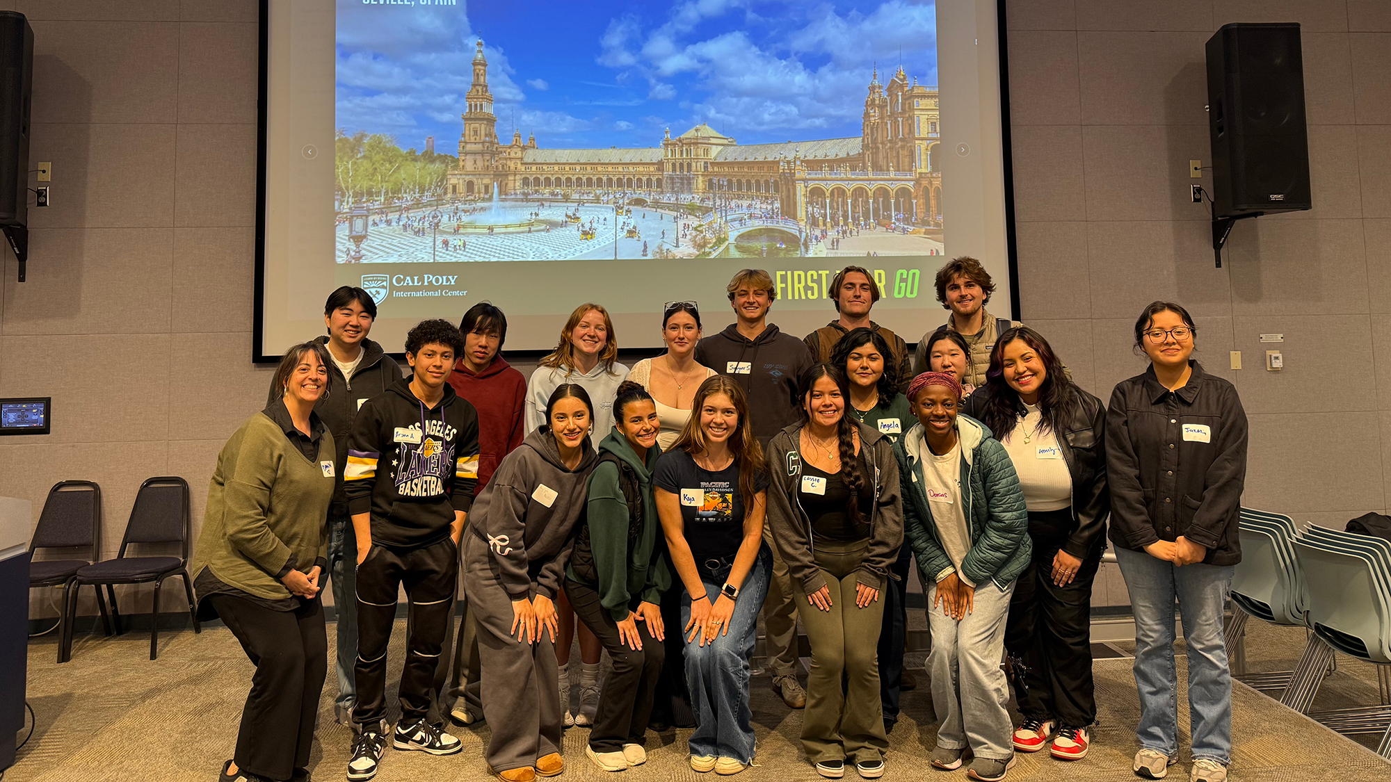 A group of Cal Poly students and staff posing together in a classroom for a group photo during the First Year Go program, with a large screen in the background displaying an image of Plaza de España in Seville, Spain and the Cal Poly International Center logo.