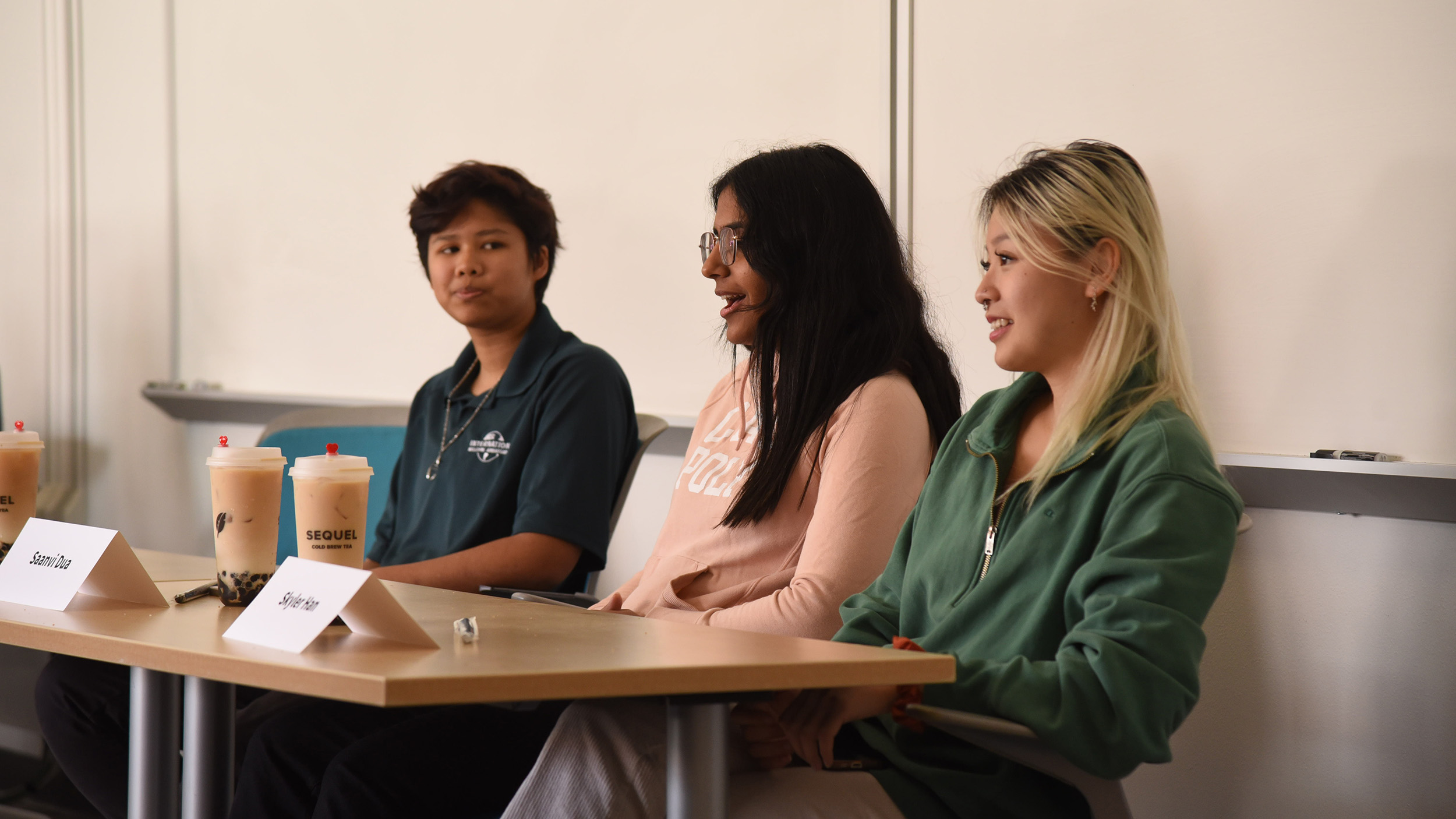 Three Cal Poly students sitting at a classroom table during a panel discussion, each with a cup of boba tea in front of them, speaking and listening attentively in front of a whiteboard.