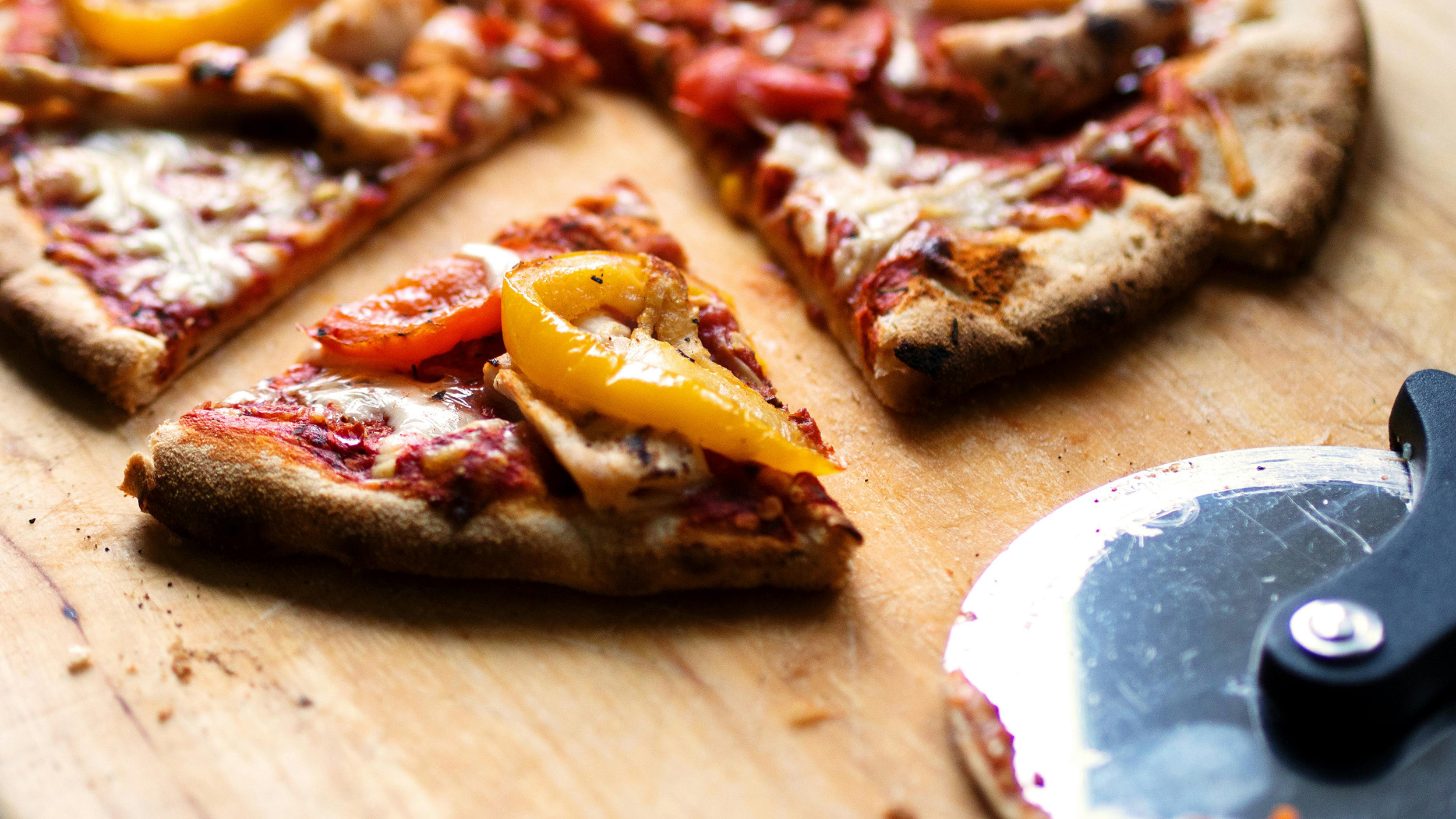 Close-up of sliced pizza with melted cheese, yellow bell peppers, and tomato sauce on a wooden board beside a pizza cutter.