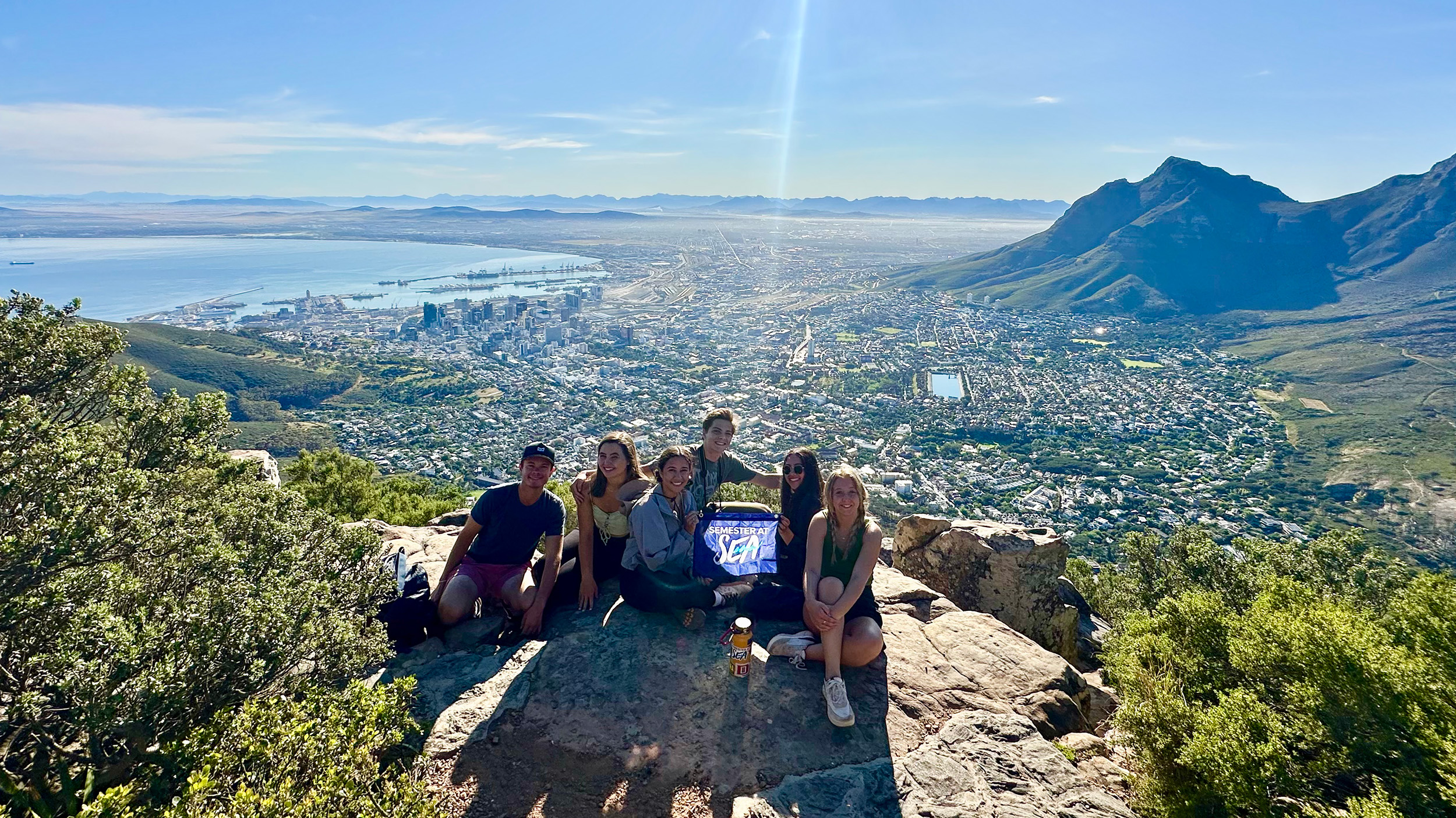 Seven students sitting on a rocky ledge atop Lion’s Head, holding a ‘Semester at Sea’ sign, with Cape Town, Table Bay, and Table Mountain in the background.