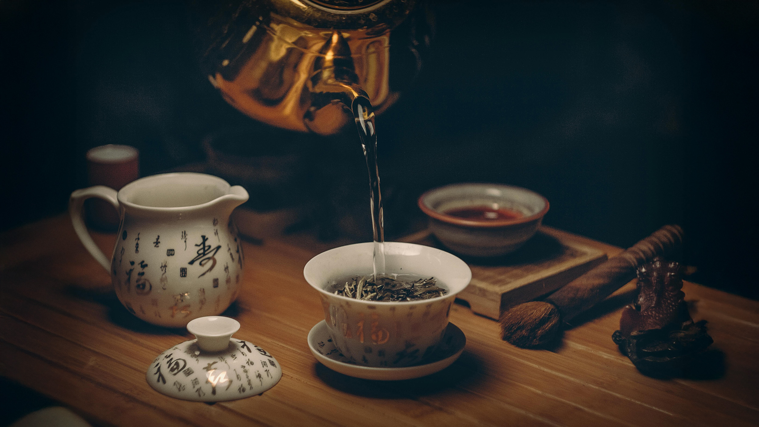 A close-up of hot water being poured from a brass teapot into a porcelain gaiwan filled with loose leaf tea during a traditional Chinese tea ceremony, surrounded by teaware on a wooden tea tray.