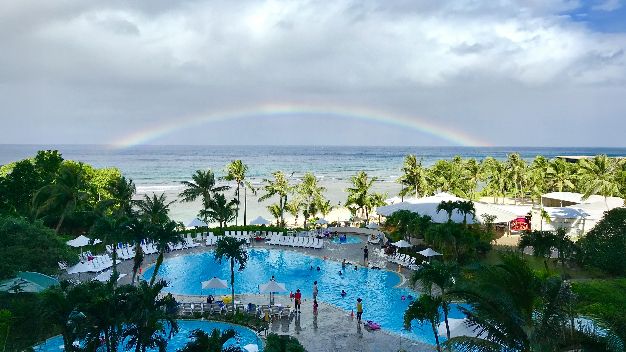 A tropical beach resort in Guam featuring a large swimming pool surrounded by palm trees and lounge chairs, with guests enjoying the pool and a vibrant rainbow arching over the ocean in the background.