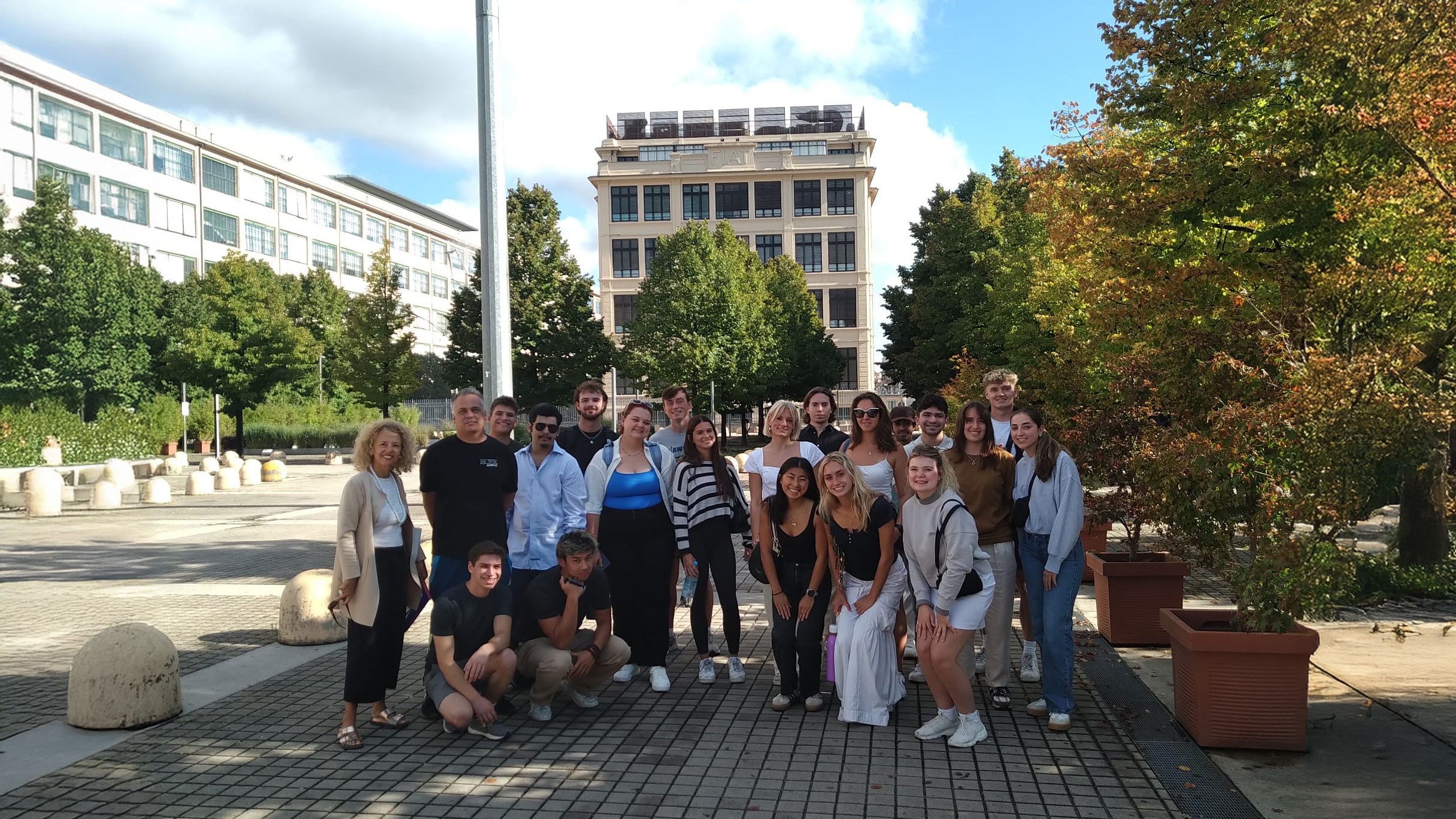 A group of study abroad students and faculty posing together outdoors on a sunny day at the USAC Torino campus in Italy, with a large academic building and trees in the background.
