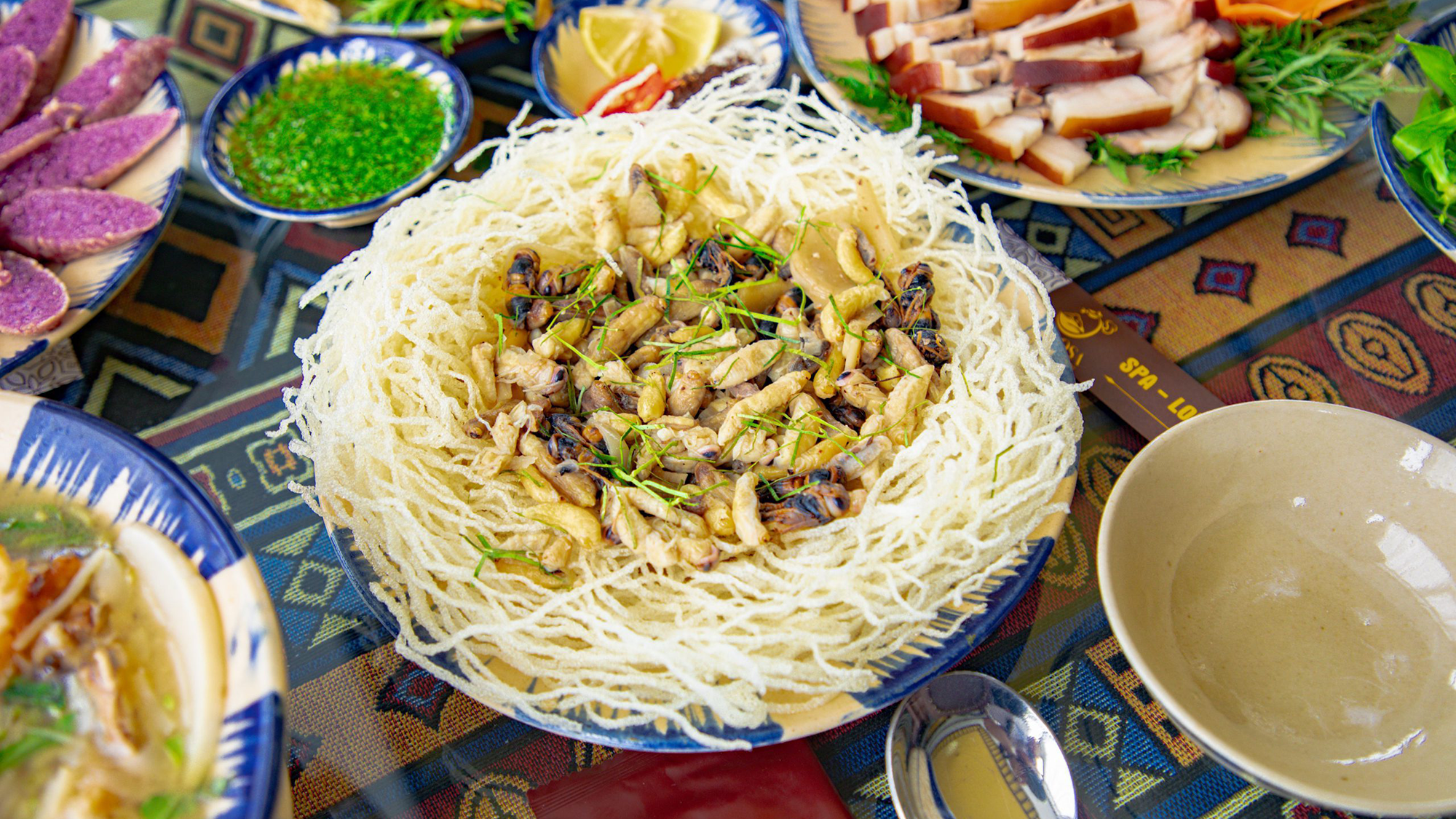 Colorful Thai meal featuring a central dish of crispy fried noodle nest filled with stir-fried mushrooms and vegetables, surrounded by assorted side dishes, sauces, and fresh ingredients on a patterned table.