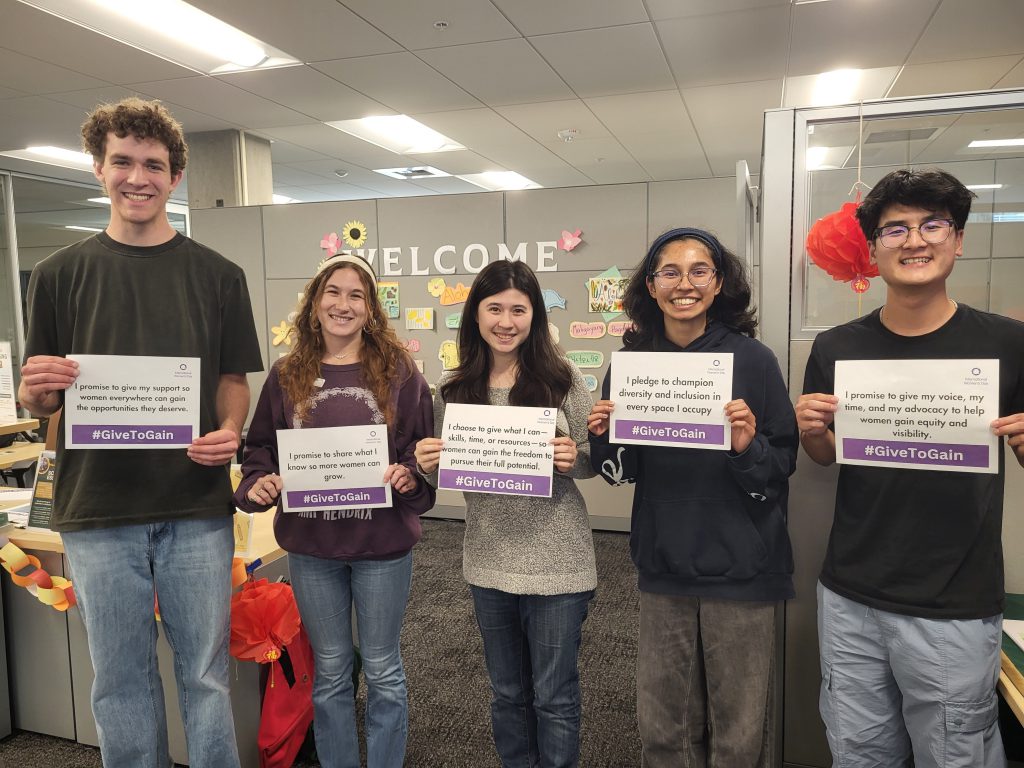Five Cal Poly students standing in an office space holding International Women’s Day #GiveToGain pledge signs promoting gender equity, diversity, and inclusion.