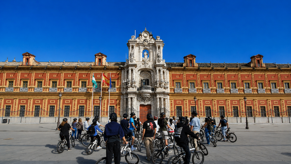 Students on a bicycle tour gathered in front of the historic Palacio de San Telmo in Seville, Spain, beneath a clear blue sky.